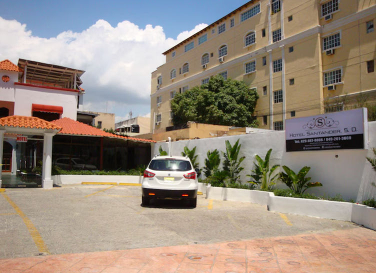Exterior facade and parking lot of Hotel Santander S.D. in Santo Domingo, featuring a white car parked in front of a sign showing the hotel name.