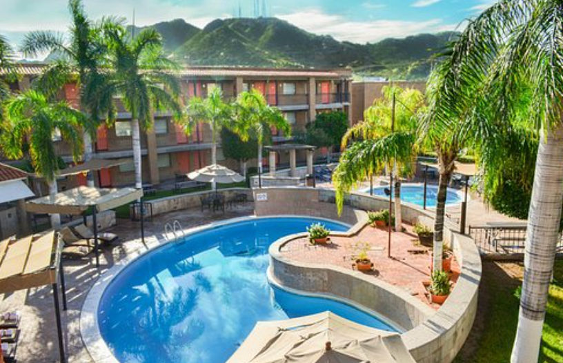 Vista aérea de la piscina curva de un hotel rodeada de palmeras, sombrillas, camastros y un edificio de habitaciones de fondo con montañas verdes bajo un cielo despejado.