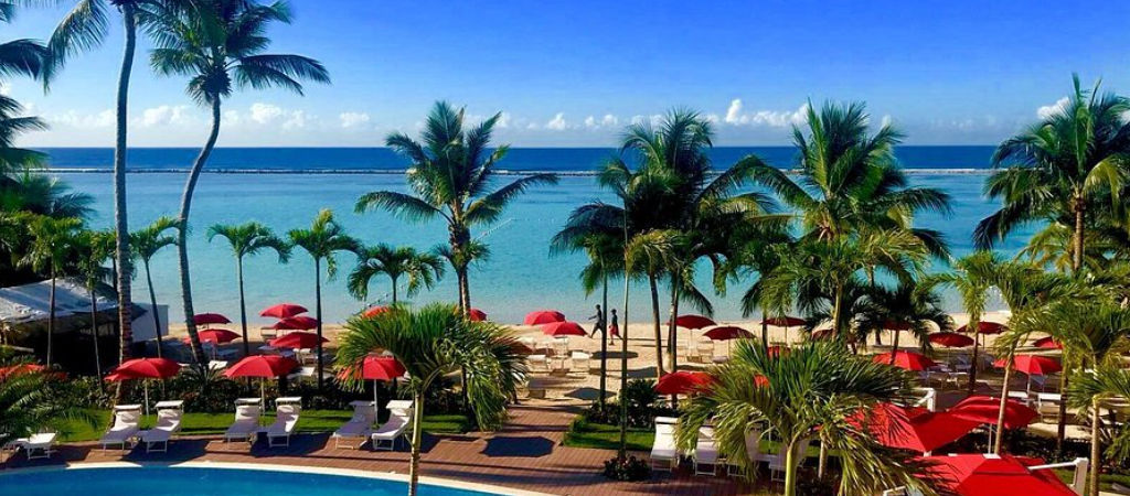 Tropical view from the swimming pool at Boca Beach Residence looking out to Boca Chica beach with palm trees, lounge chairs, and red umbrellas facing the blue Caribbean Sea.