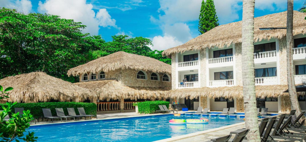 Piscina al aire libre rodeada de tumbonas y edificios de estilo tropical con techos de palma bajo un cielo azul