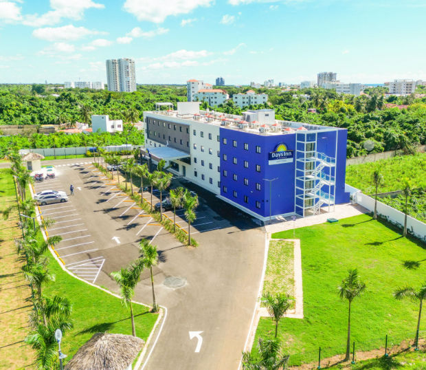 Vista aérea del hotel Days Inn By Wyndham Juan Dolio, mostrando la fachada moderna del edificio azul y blanco, el estacionamiento y las áreas verdes circundantes.