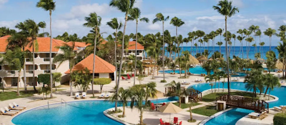 Panoramic view of the large outdoor swimming pools at the Jewel Palm Beach hotel in Punta Cana, surrounded by tropical palm trees, colonial tiled-roof buildings, and the beach in the background.