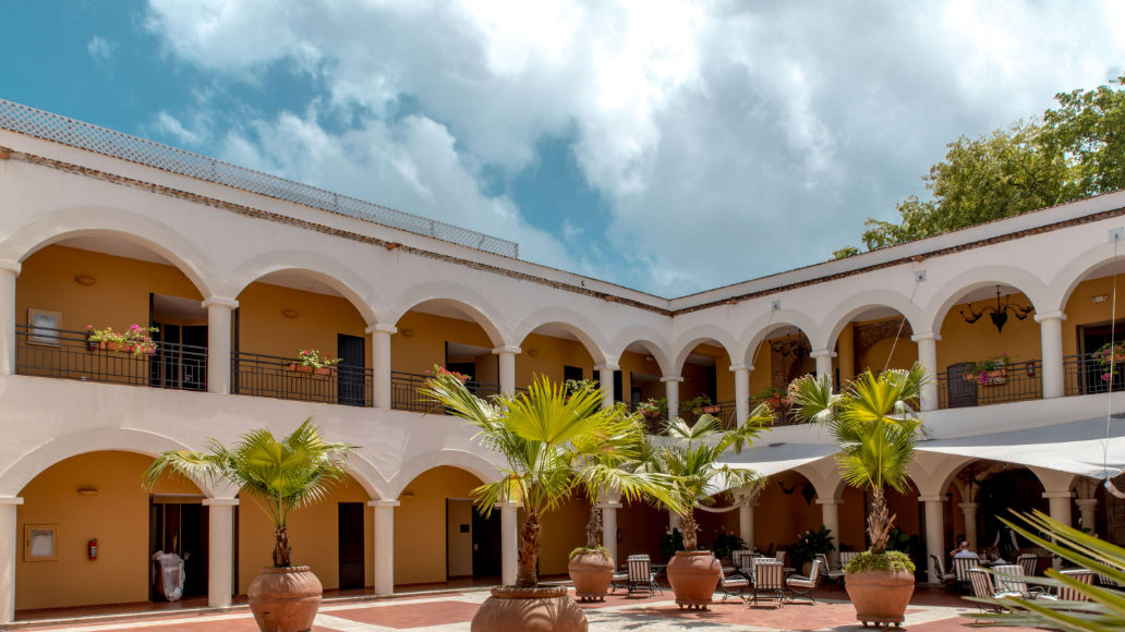 Colonial-style interior courtyard featuring two-tiered archways, palm trees in large clay pots, outdoor seating tables, and a partly cloudy sky.
