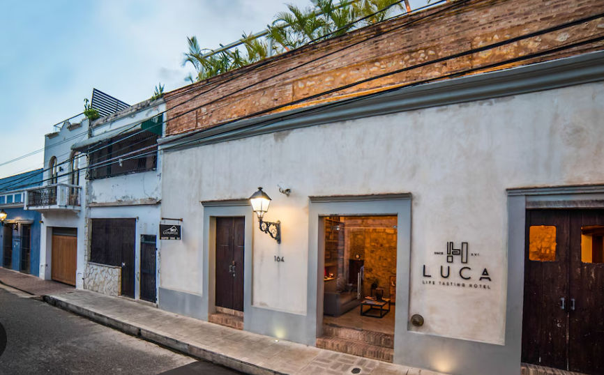 Exterior facade of the Luca Life Tasting Hotel in the Colonial Zone of Santo Domingo, showcasing its historic colonial-style architecture with white stucco walls, dark wood doors, a classic street lamp, and plants on the rooftop terrace.