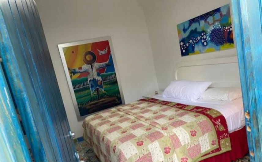 Cozy and simple bedroom at Chao Mama Hotel featuring a double bed with white linens, a bedside lamp on a glass table, blue curtains, and a decorative sailboat painting on the wall.