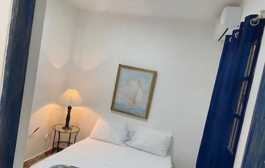 Cozy and simple bedroom at Chao Mama Hotel featuring a double bed with white linens, a bedside lamp on a glass table, blue curtains, and a decorative sailboat painting on the wall.