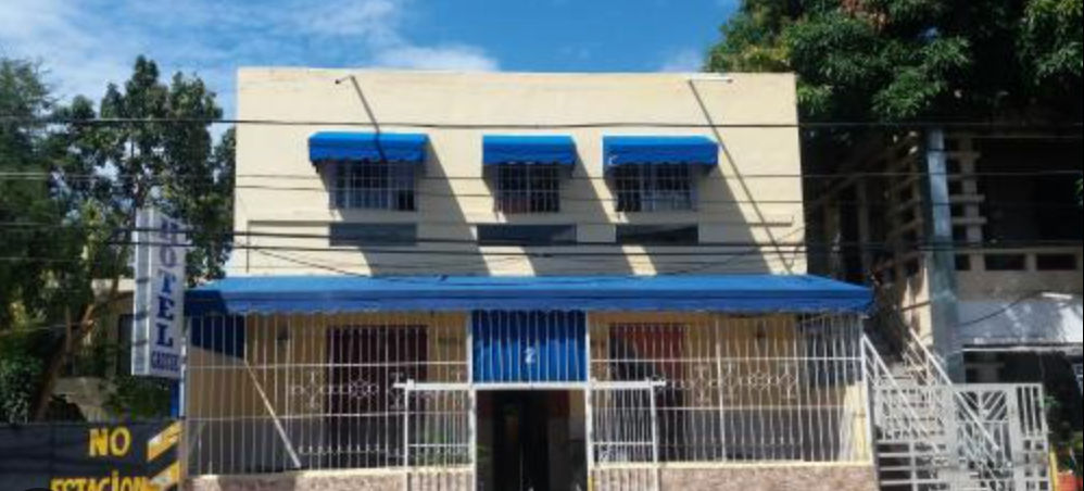 Fachada exterior del Hotel Gazcue mostrando un edificio de dos pisos color crema con toldos azules y rejas de metal en la entrada.