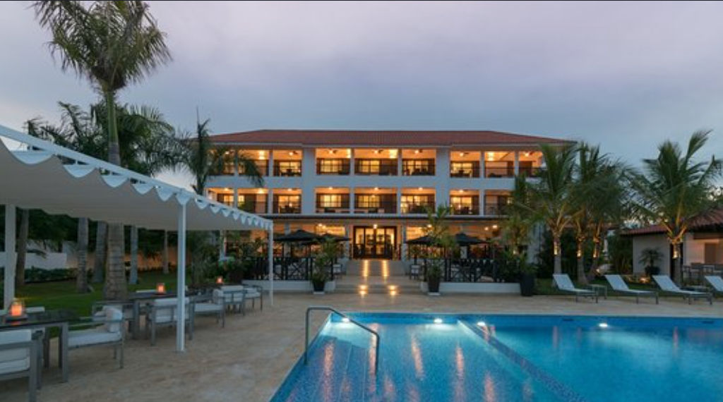 Piscina iluminada por la noche frente a la fachada principal del Hotel Casa Hemingway, un edificio de tres niveles de estilo boutique rodeado de palmeras en Juan Dolio.