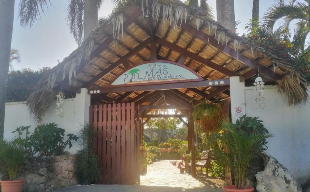 Vista exterior de una encantadora casa tropical con techo inclinado verde, porche y jardín exuberante con palmeras y flores bajo un cielo azul con un arcoíris