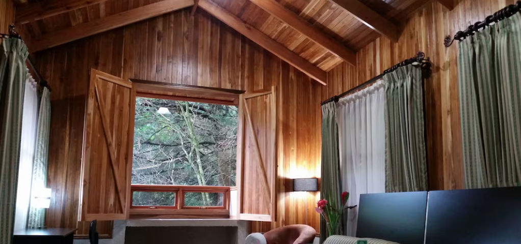 A rustic wooden cabin with a stone chimney nestled in a lush green forest with purple flowers in the foreground / Cabaña rústica de madera con chimenea de piedra en un frondoso bosque verde con flores moradas en primer plano