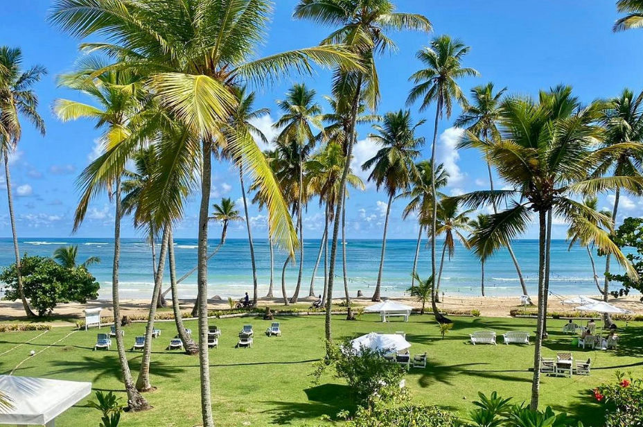 Vista exterior de un hotel boutique tropical con arquitectura colonial blanca, un cenador tradicional, una gran palmera y jardines verdes bajo un cielo azul