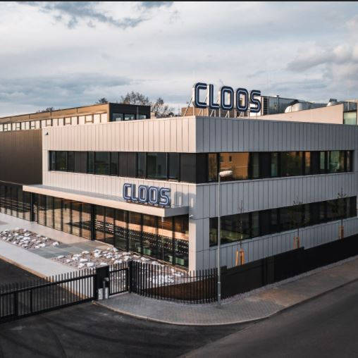 Modern factory building with "Cloos" signage and spacious parking lot, taken under a cloudy sky.