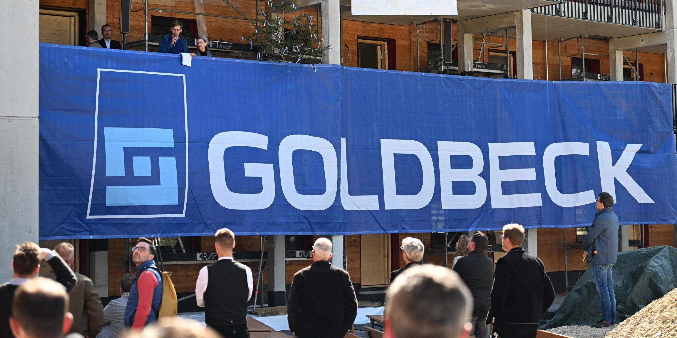 Gebäudeeinweihung mit großem Goldbeck-Banner, Menschenmenge schaut zu, im Hintergrund ein Bauprojekt mit 100-Jahre-Feier.