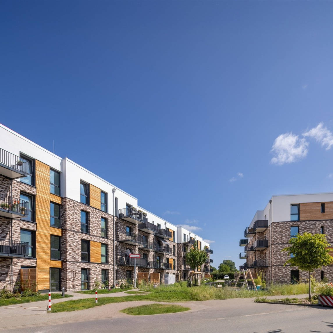 Modern residential complex with balconies and parking spaces, surrounded by green spaces, under a blue sky.