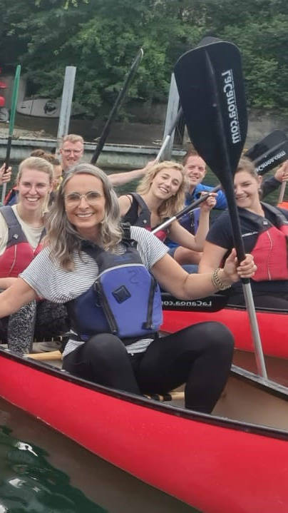 Gruppe von Menschen beim Kanufahren auf einem Fluss, lächelnd und teils mit Paddeln in der Hand, im Hintergrund Bäume und ein Gebäude.