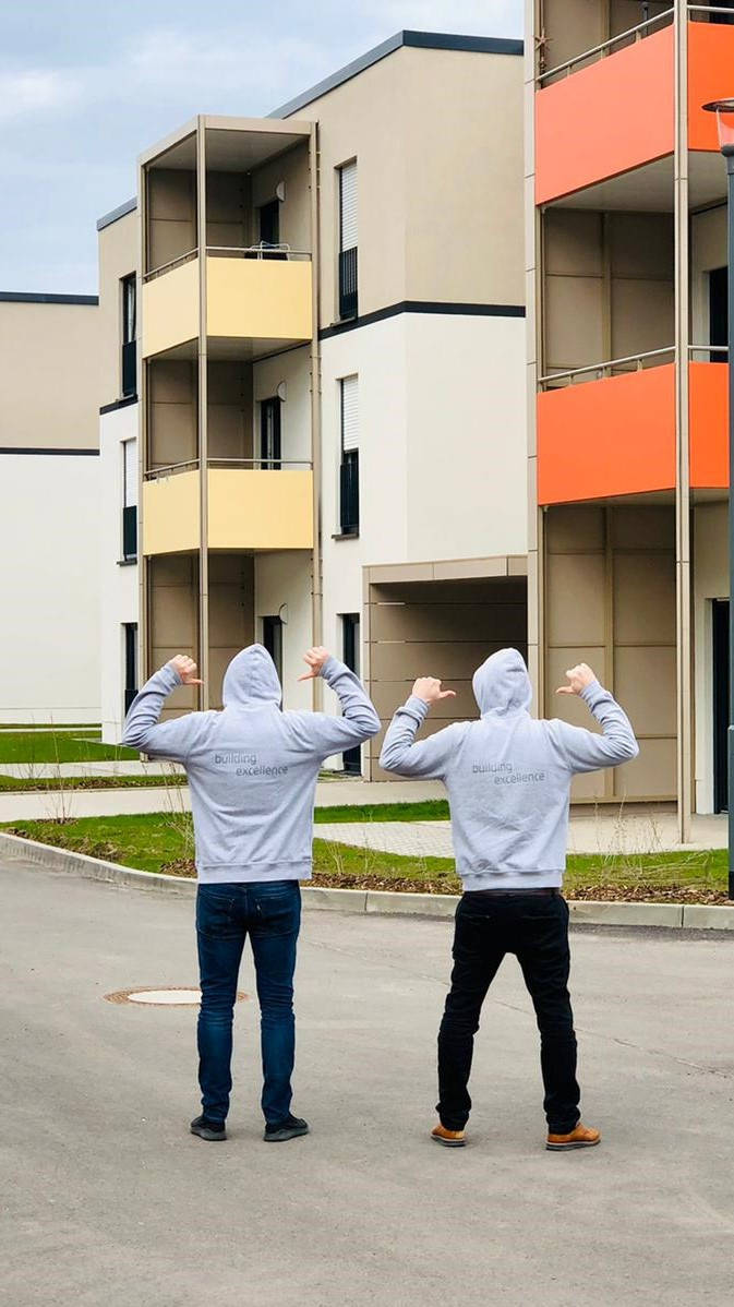 Zwei Personen in grauen Hoodies stehen vor modernen Wohngebäuden und zeigen mit den Daumen auf ihren Rücken.