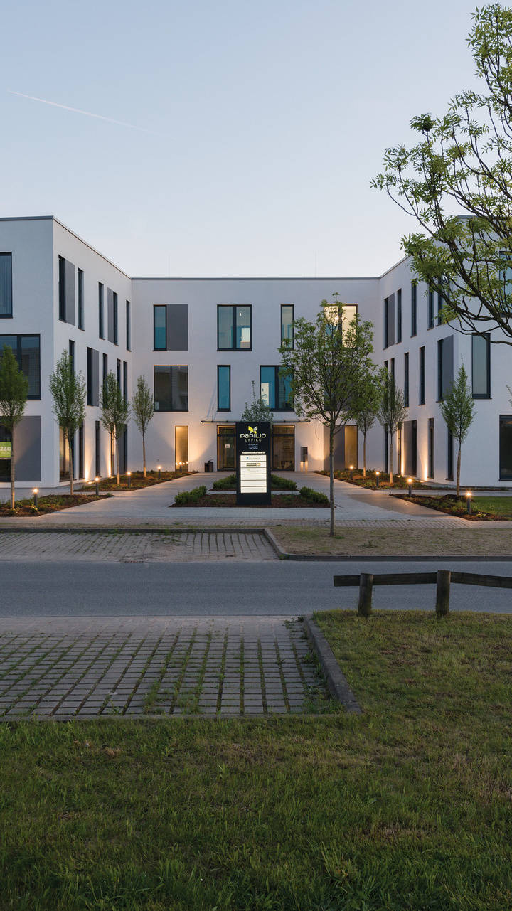 Modernes Bürogebäude mit weißer Fassade und großen Fenstern, umgeben von gepflegten Grünflächen und Bäumen bei Dämmerung.