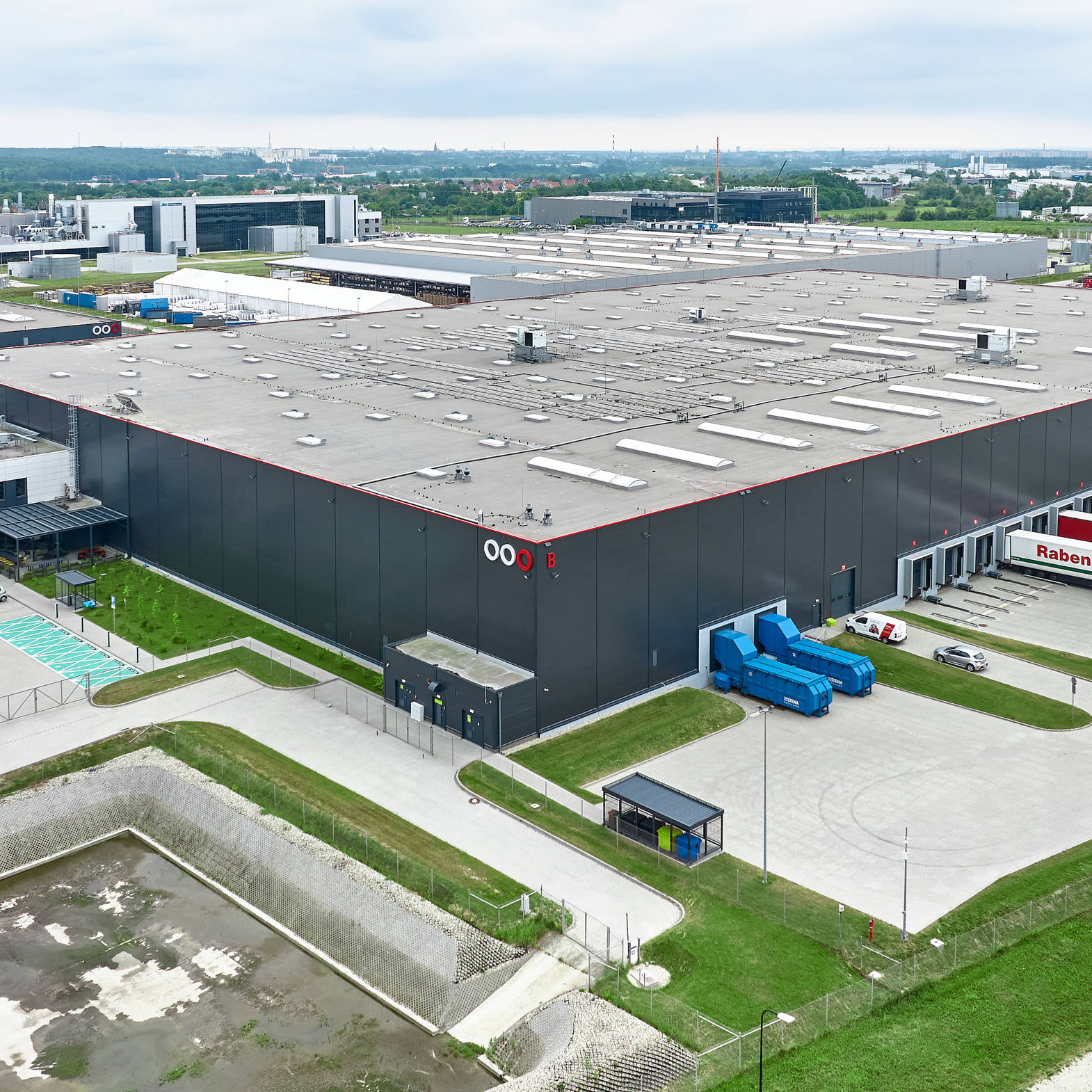 Zdjęcie przedstawia duży, nowoczesny magazyn przemysłowy widziany z lotu ptaka. Z lewej strony znajduje się parking z zaparkowanymi samochodami, natomiast z prawej strony widać kilka ciężarówek zaparkowanych przy rampach. Budynek jest otoczony zielenią, a na horyzoncie widać inne budynki przemysłowe oraz tereny zielone.