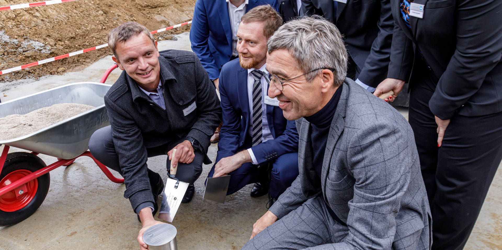 Männer in Anzügen führen auf einer Baustelle eine symbolische Grundsteinlegung mit Metallzylinder und Kelle durch.