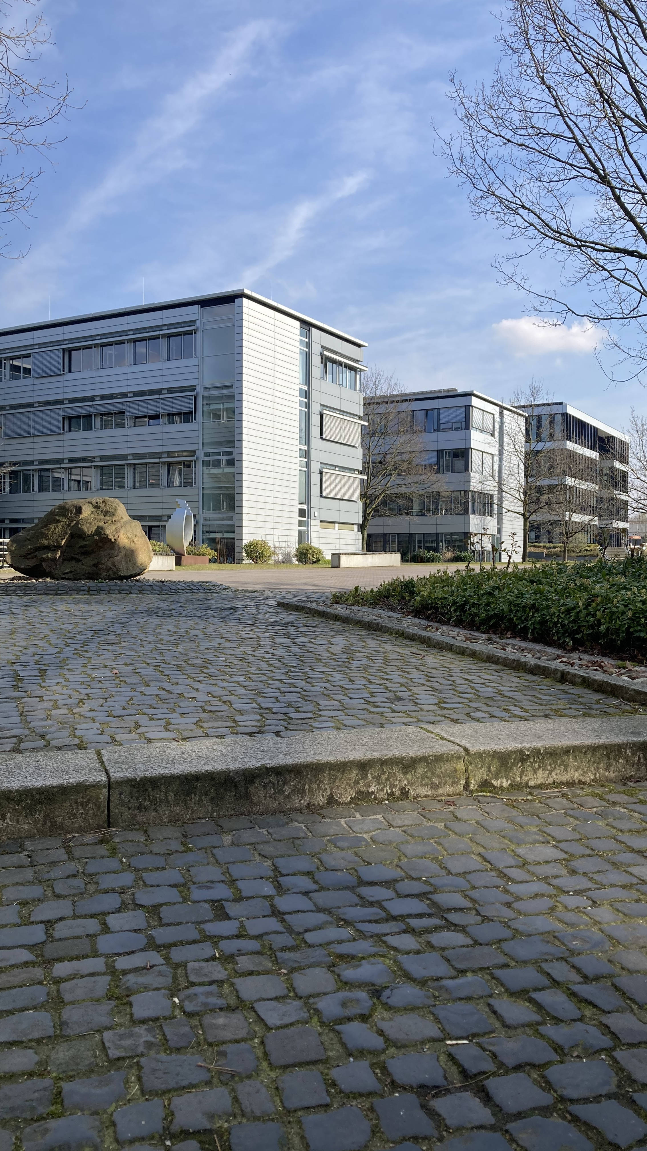 Modernes Bürogebäude an einem klaren, sonnigen Tag, umgeben von kahlen Bäumen und einem gepflasterten Weg im Vordergrund.
