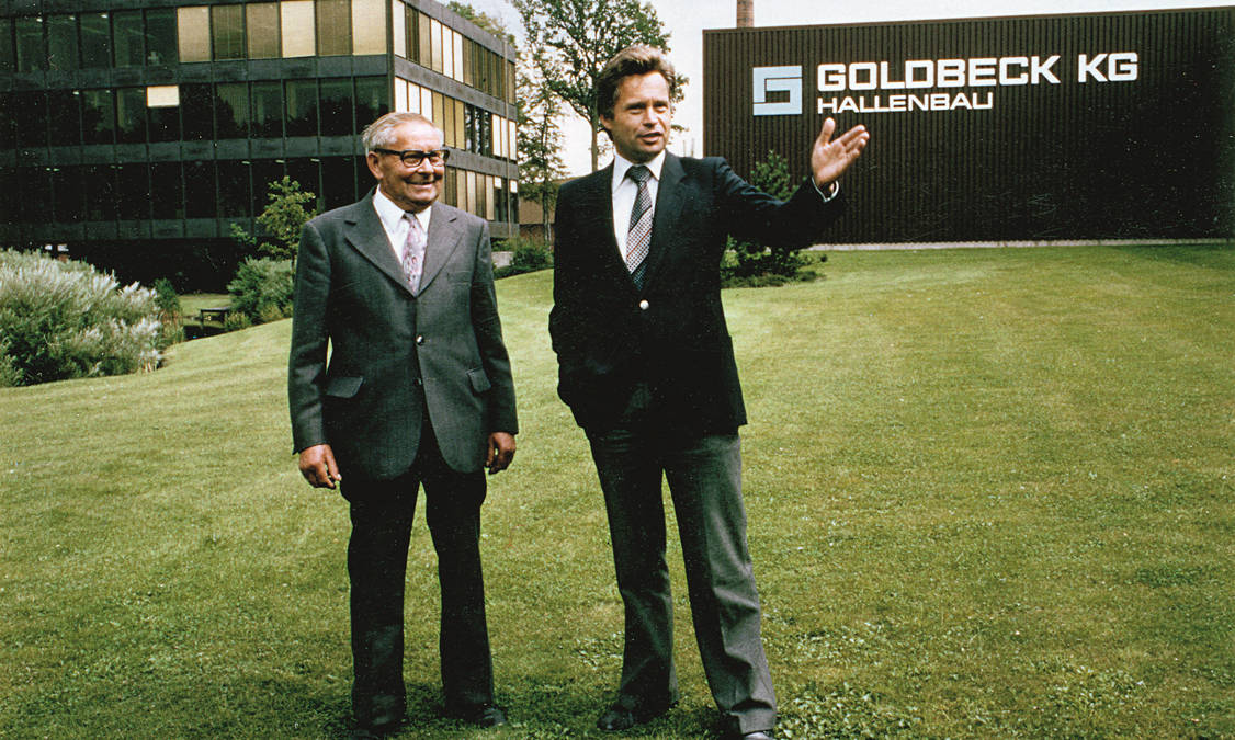 Two men in suits are standing on a large lawn. In the background is an office building and an industrial building.