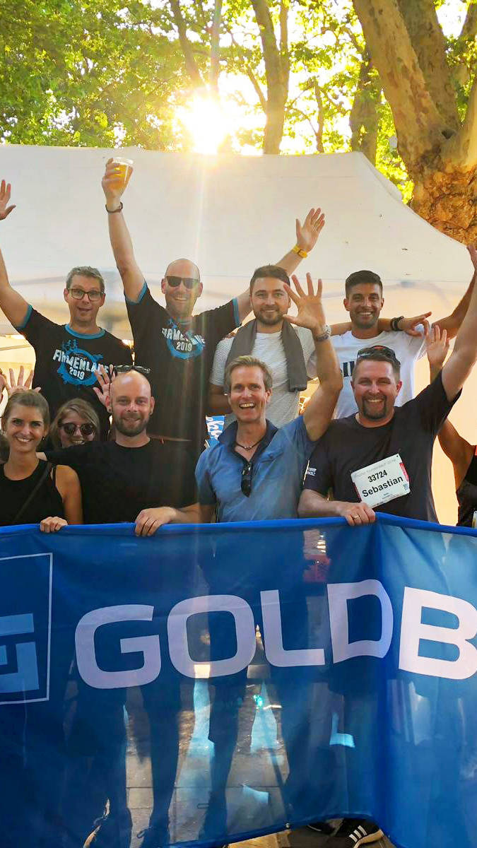 Gruppe von Teilnehmern beim Firmenlauf 2019, die jubelnd ein "Goldbeck"-Banner halten, mit Bäumen und Sonnenschein im Hintergrund.