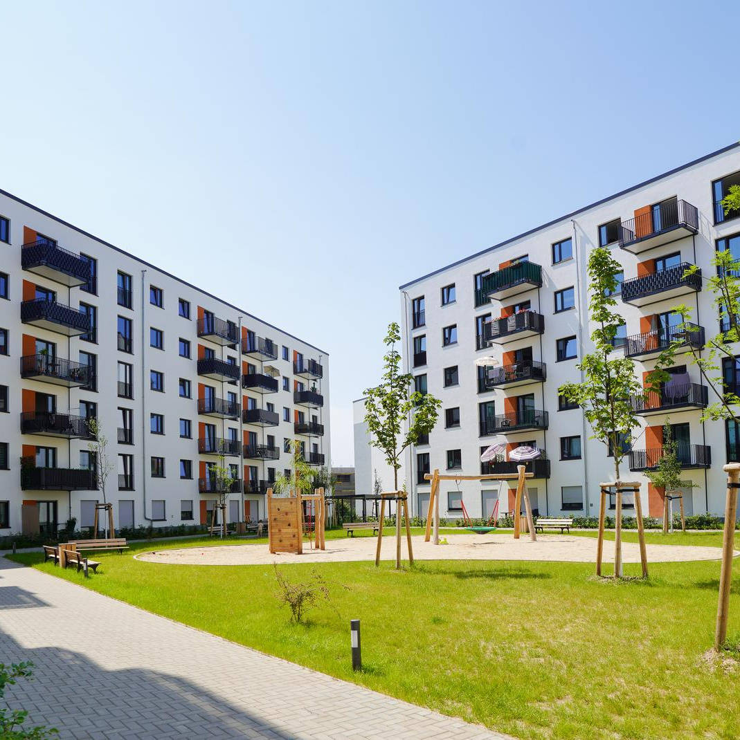 Multi-storey residential buildings around an inner courtyard with a playground and young trees, sunny sky in the background.