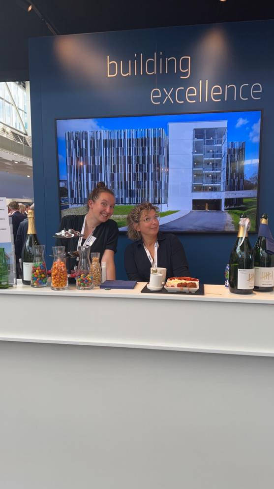 Messestand mit zwei Frauen hinter einer Tresen, dahinter ein großes Bild eines modernen Gebäudes und der Text "Building Excellence". Auf dem Tresen stehen Champagnerflaschen und Snacks.