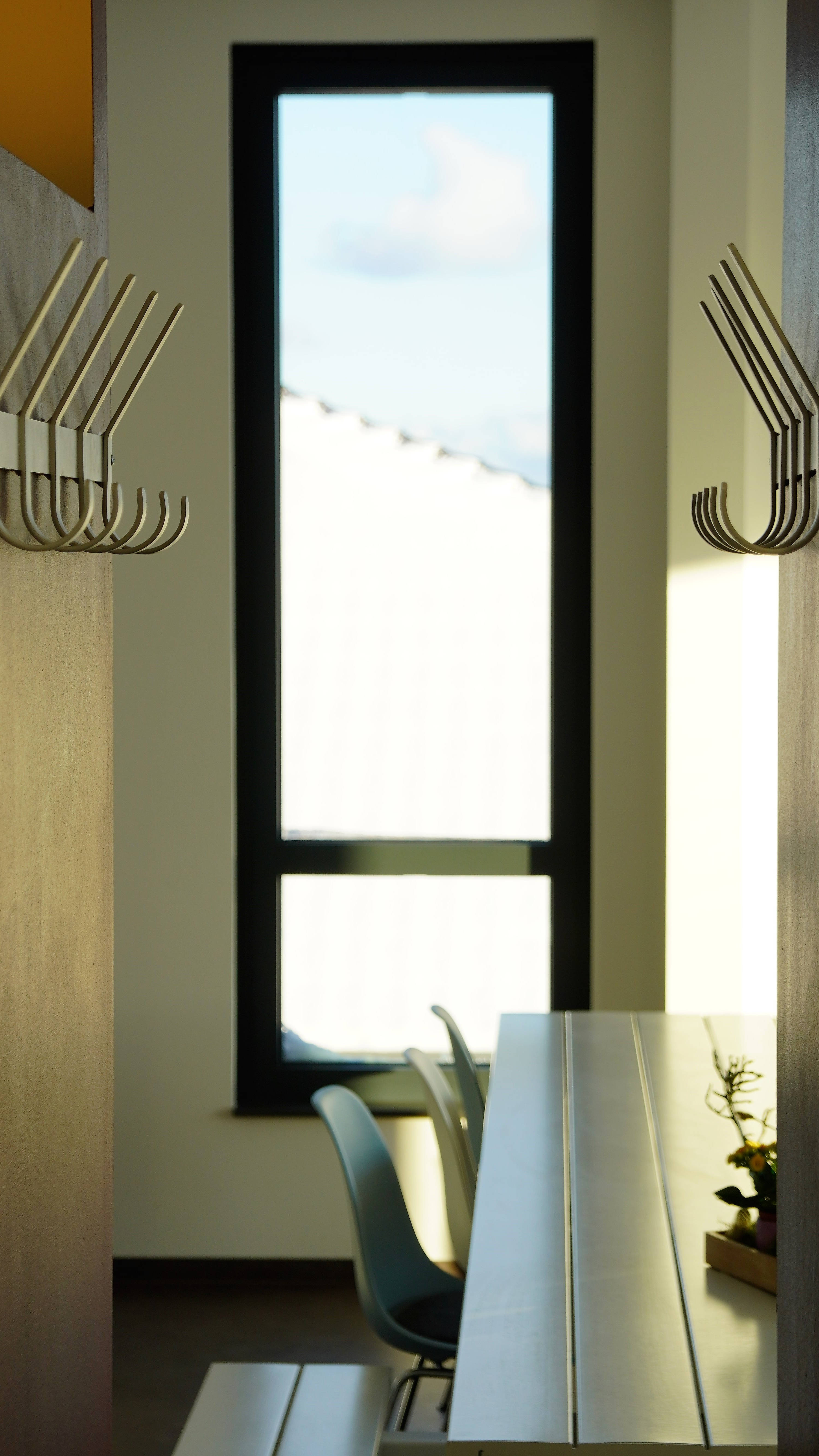 Modern gestalteter Innenraum mit Kleiderhaken, einem Tisch und Stühlen vor einem großen Fenster, durch das Licht einfällt.