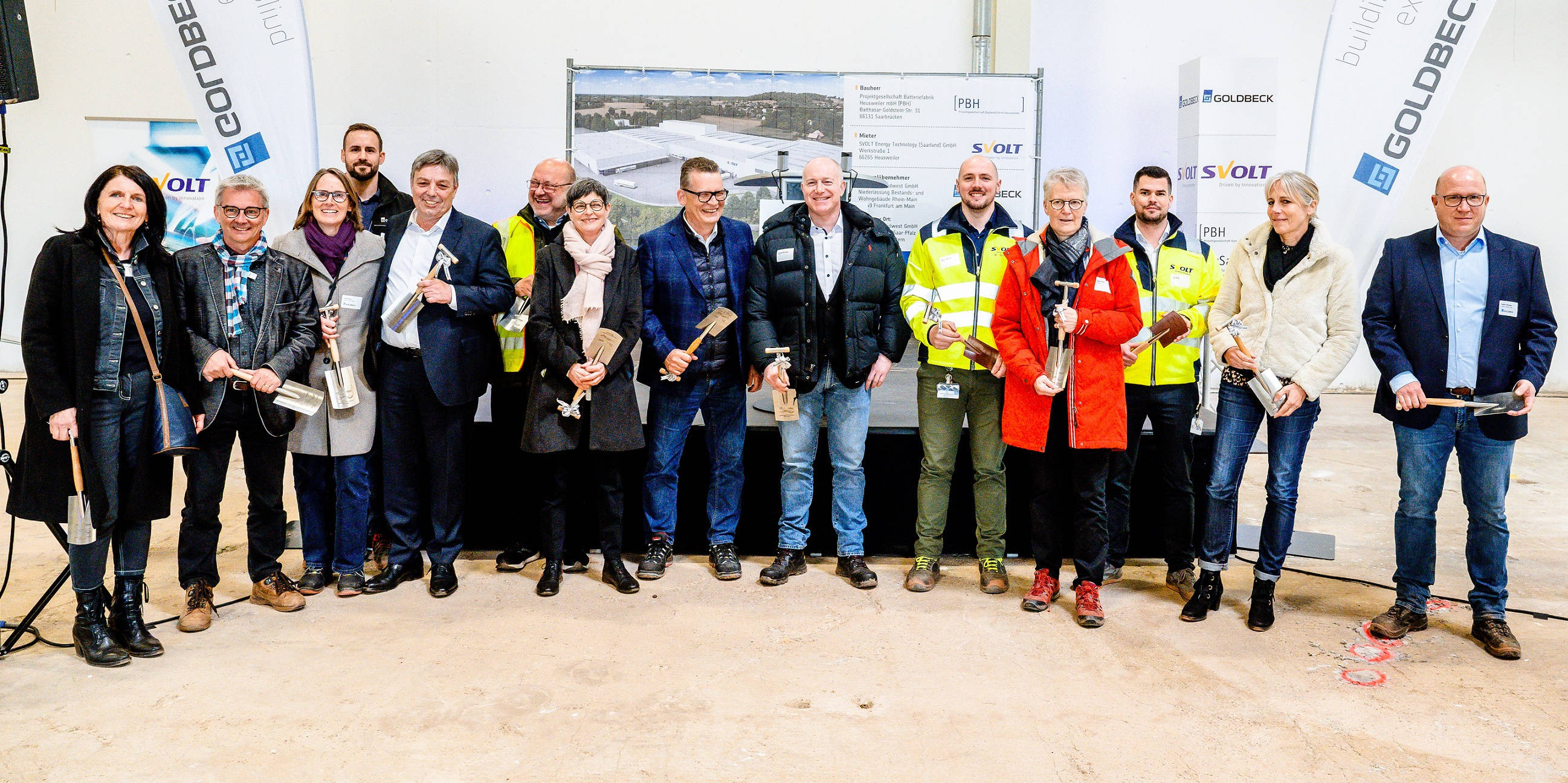 Gruppe von Personen in einem Industriegebäude hält Spaten und feiert den Spatenstich eines Bauprojekts, umgeben von Firmenlogos.