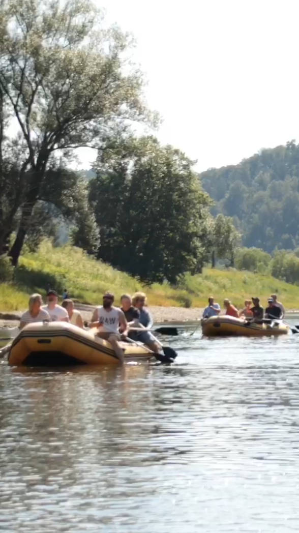 Mehrere Gruppen von Menschen in Schlauchbooten fahren auf einem ruhigen Fluss, umgeben von bewaldeten Hügeln und Bäumen.