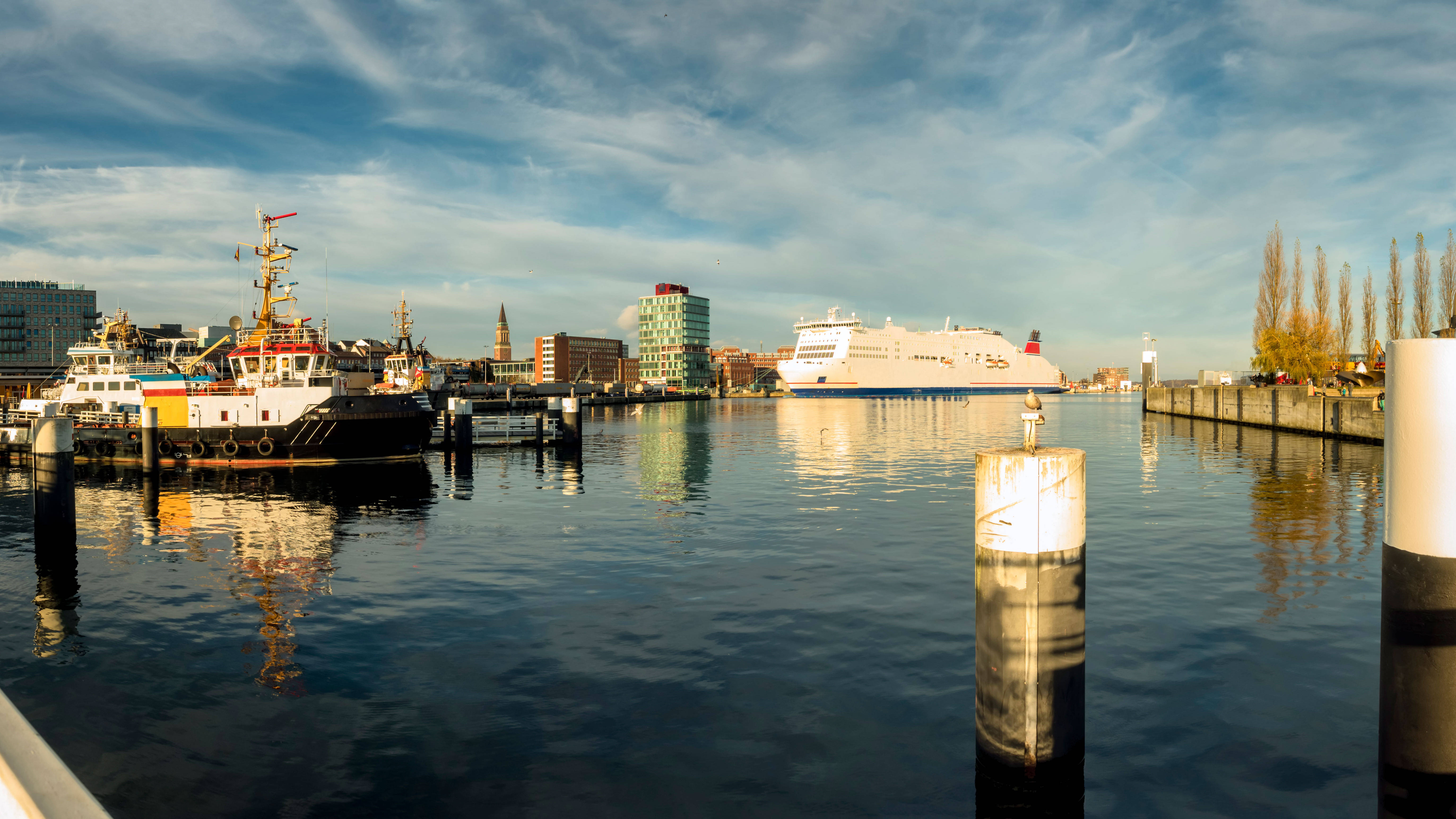 Panorama eines Hafens, mit einem großen Fährschiff und mehreren Booten im Vordergrund, umgeben von modernen Gebäuden und einem klaren blauen Himmel im Hintergrund.