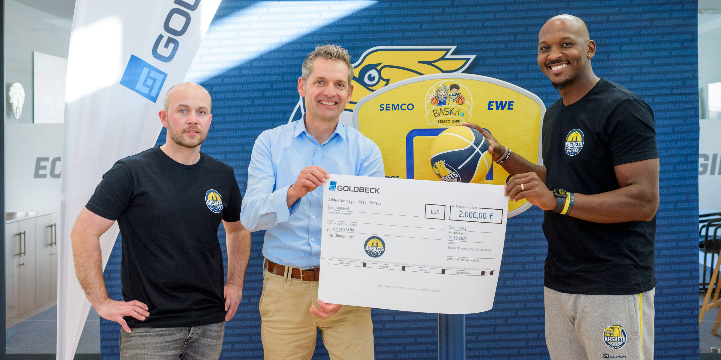 Eine Gruppe von drei Männern, die einen großen Scheck über 2000 Euro hält, vor einem Banner mit Basketball-Motiven, vermutlich im Rahmen einer Sponsorenveranstaltung.