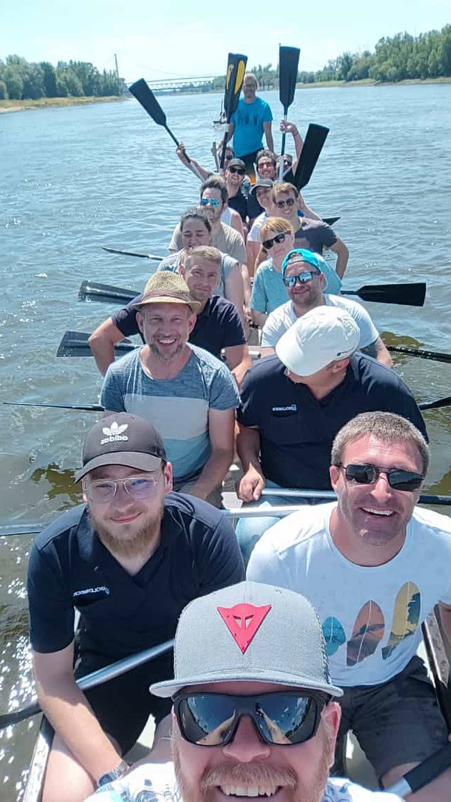 Gruppe von Menschen beim Rudern auf einem Fluss an einem sonnigen Tag. Im Hintergrund sind Bäume und eine Brücke zu sehen.