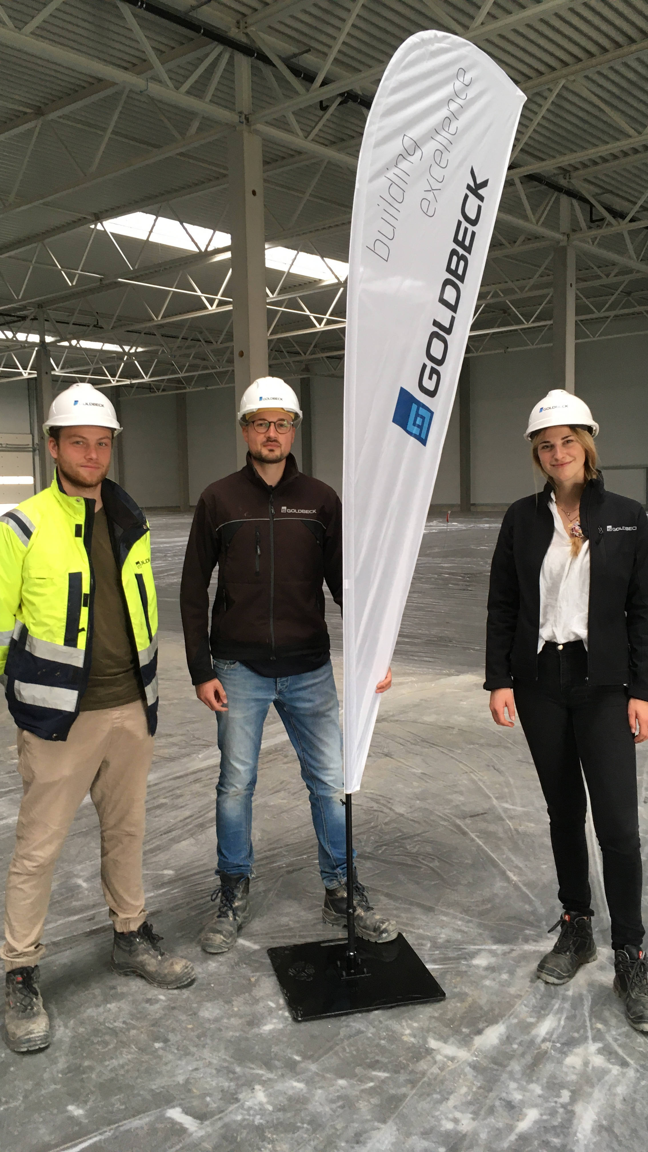 Drei Personen mit Helmen in einem leeren Industriegebäude stehen neben einem Goldbeck-Werbeschild.