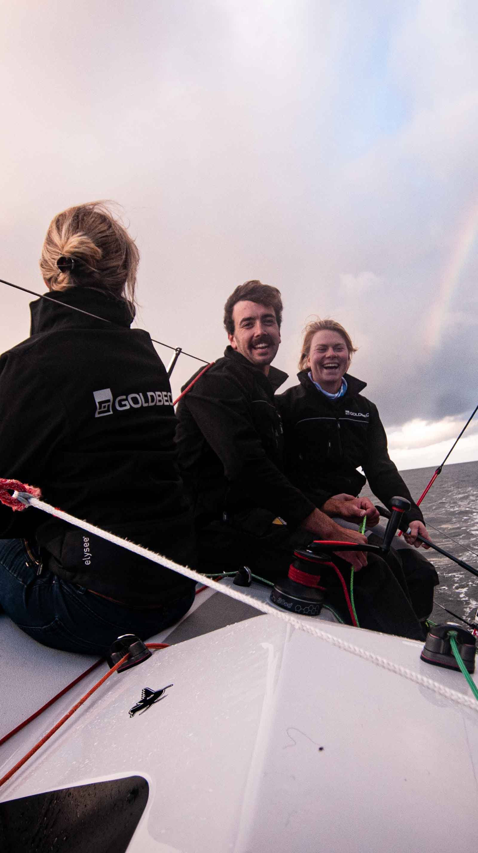 Drei Personen segeln bei leicht bewölktem Himmel und Regenbogen auf einem offenen Gewässer, lachen und genießen die Fahrt.