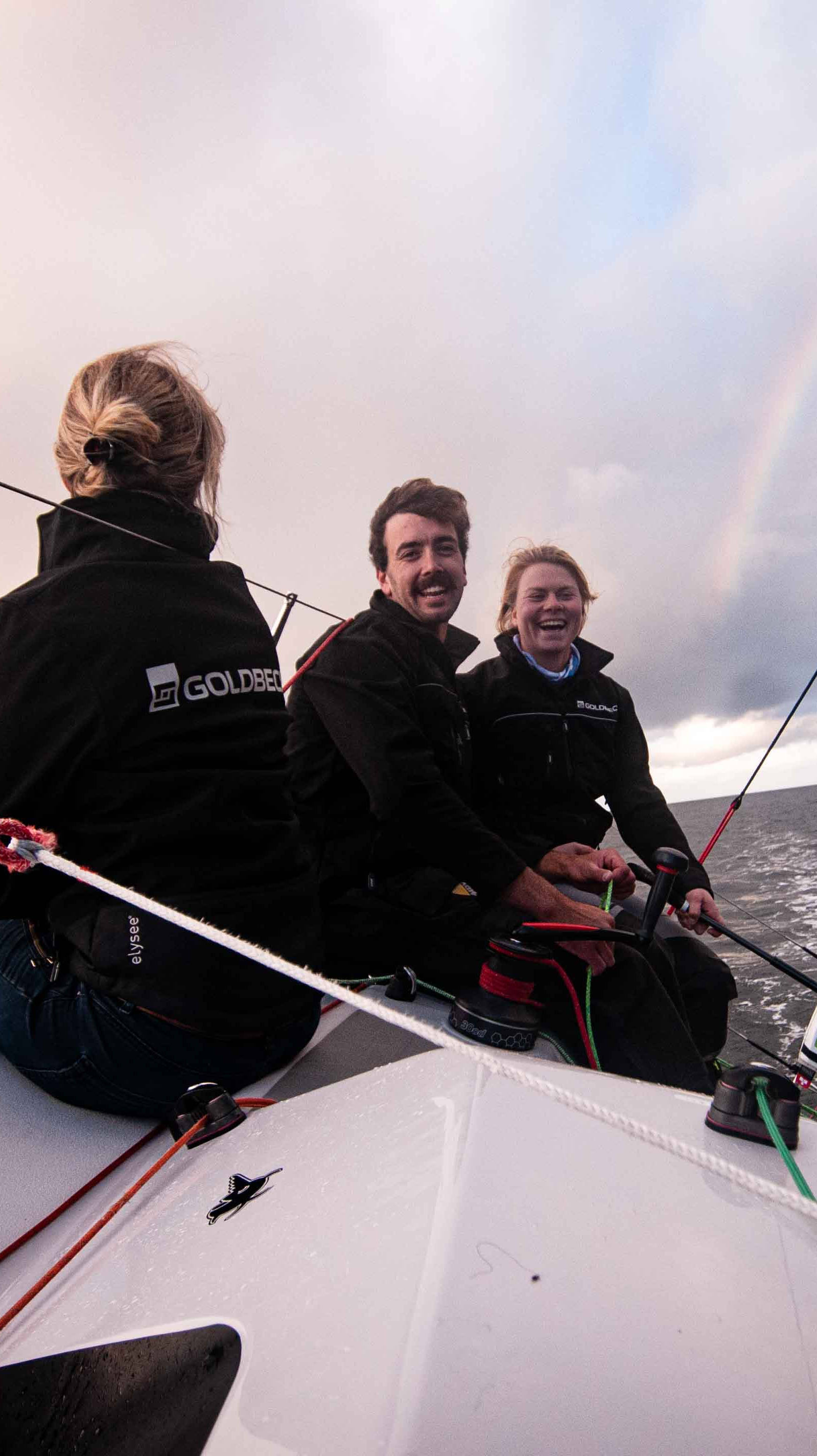 Drei Personen segeln bei leicht bewölktem Himmel und Regenbogen auf einem offenen Gewässer, lachen und genießen die Fahrt.