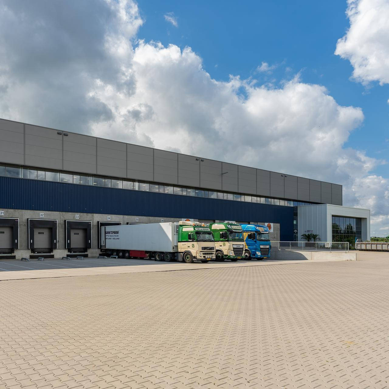 Een groot magazijn met meerdere laad- en losdeuren. Voor het gebouw staan verschillende vrachtwagens, waarvan twee groene en een blauwe. De lucht is gedeeltelijk bewolkt met hier en daar blauwe plekken.