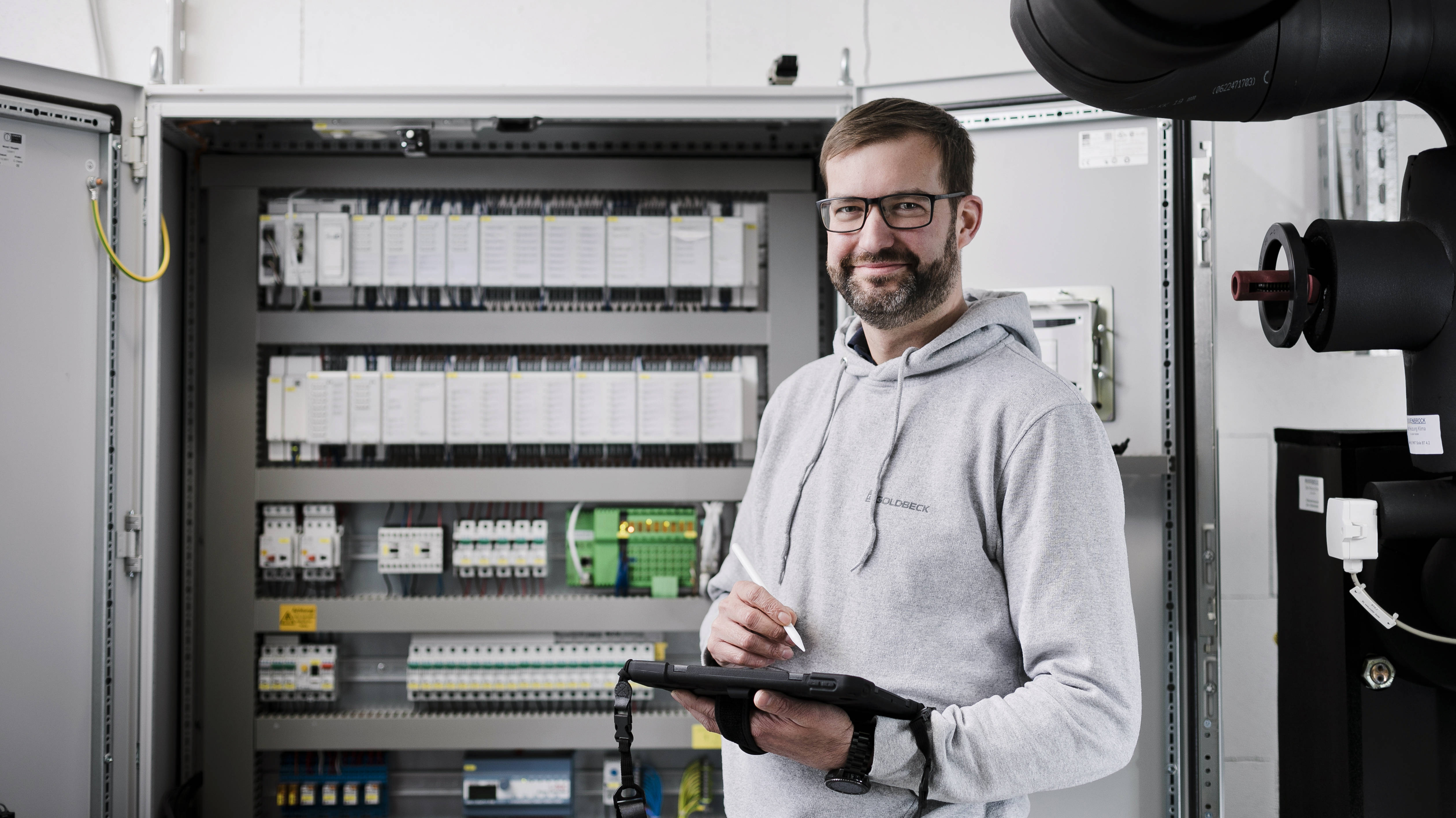 A technician in a gray hoodie stands with a tablet in front of an open control cabinet in an industrial space.