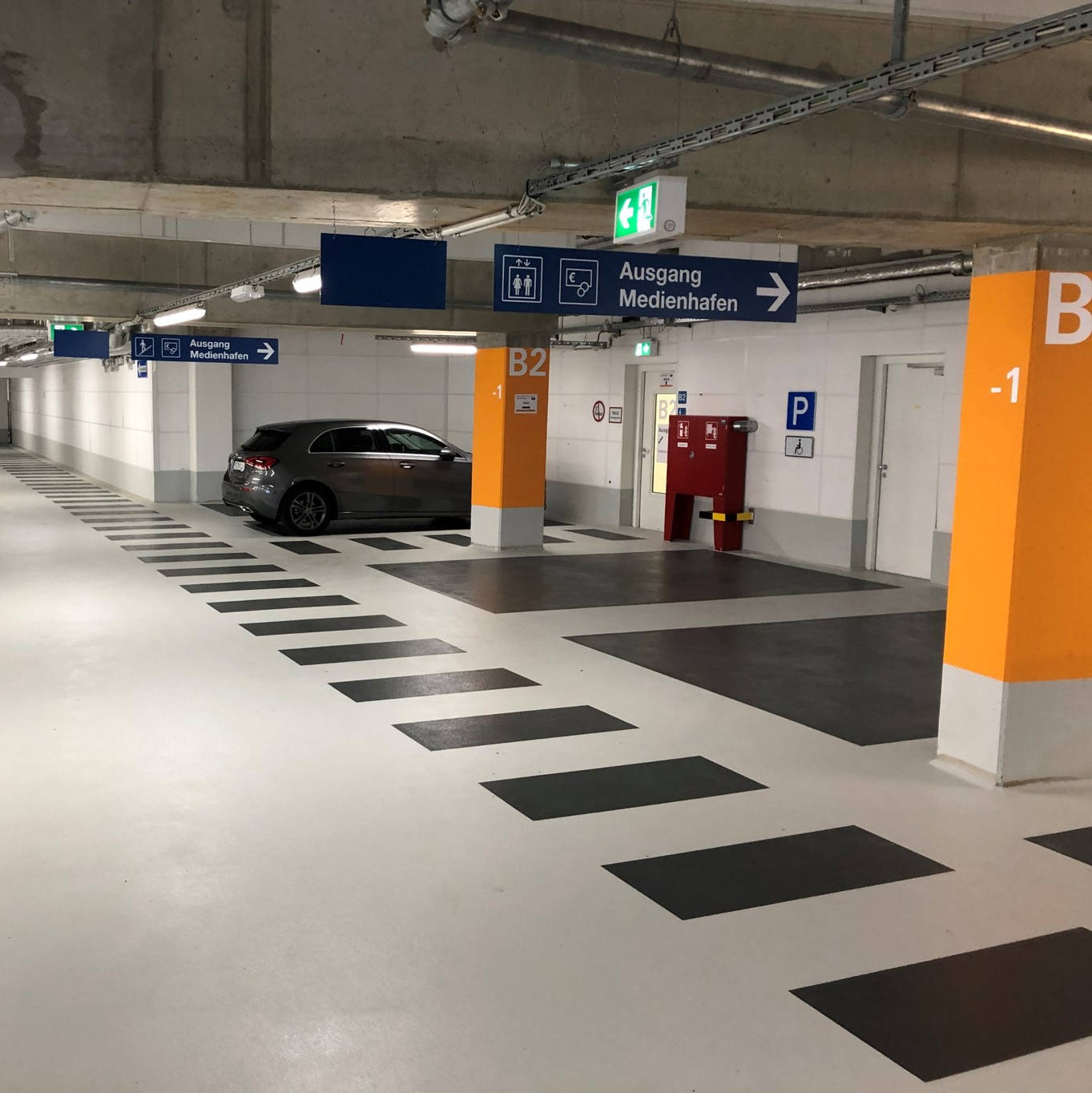 Parkhaus Innenansicht mit markierten Stellplätzen, Hinweisschildern zum Ausgang "Medienhafen" und mehreren geparkten Autos.