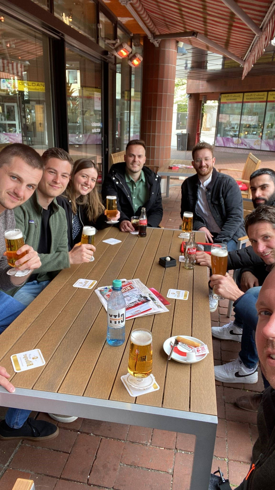 Gruppe von acht Personen sitzt in einem Café im Freien, trinkt Bier und lächelt in die Kamera.