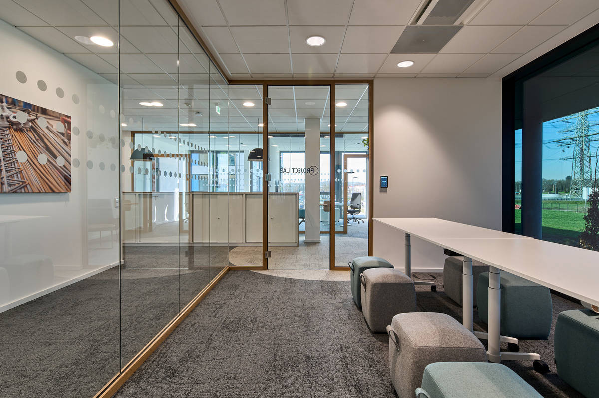 Modern conference room with glass partition wall, large table and upholstered stools, view of office area through glass doors in the background.