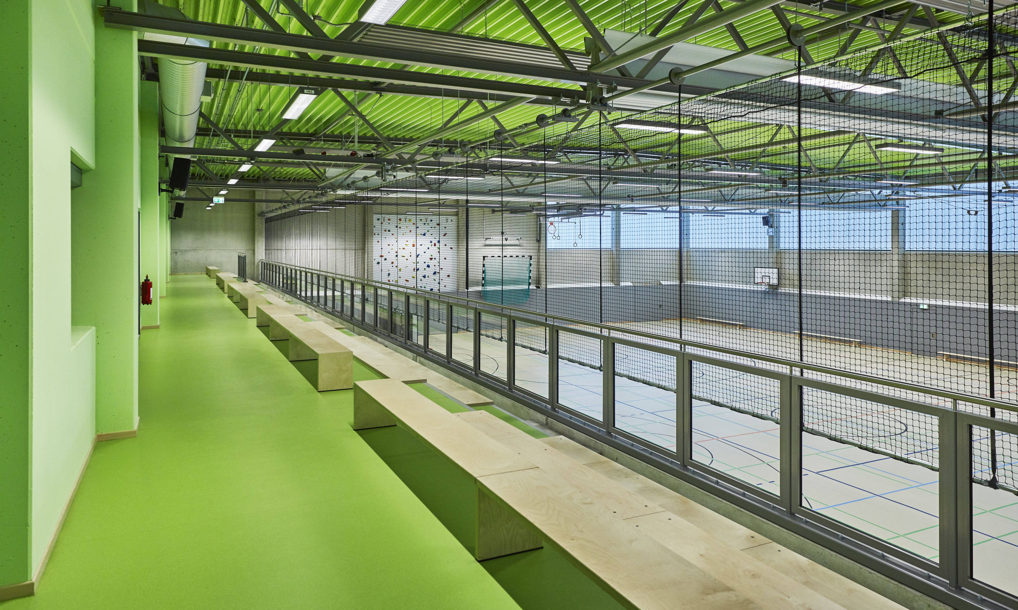 A green spectator stand overlooking a modern sports hall with a climbing wall and a basketball hoop.