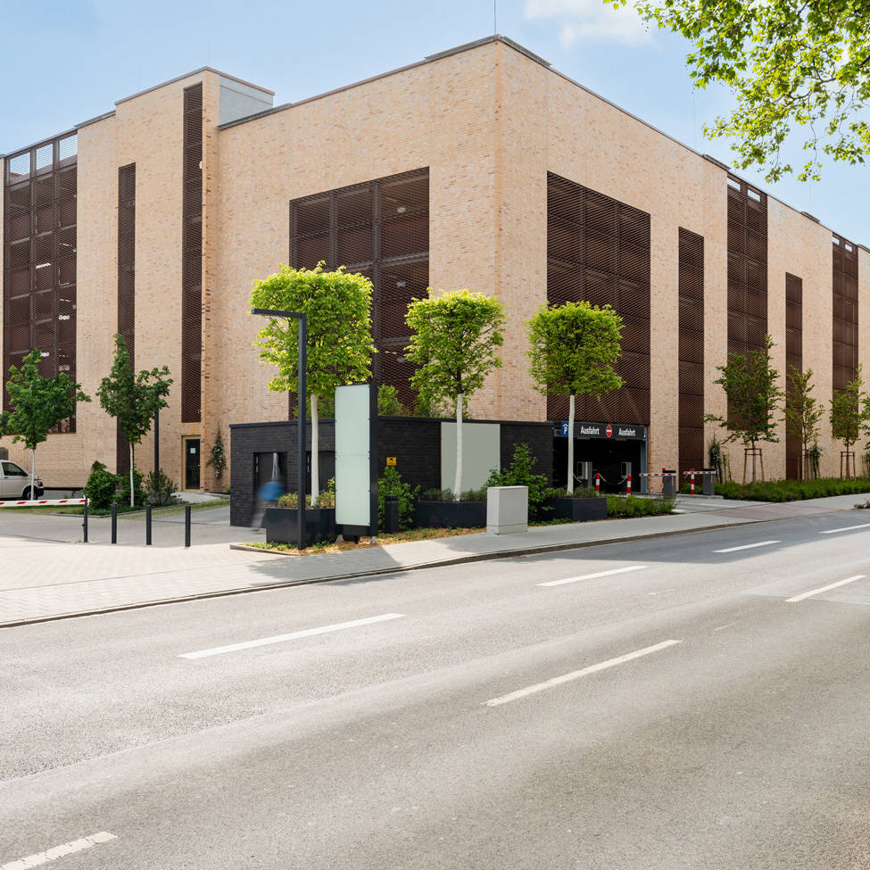 Modernes Parkhaus aus hellem Backstein mit begrünten Außenanlagen und Zufahrt an einer städtischen Straße.