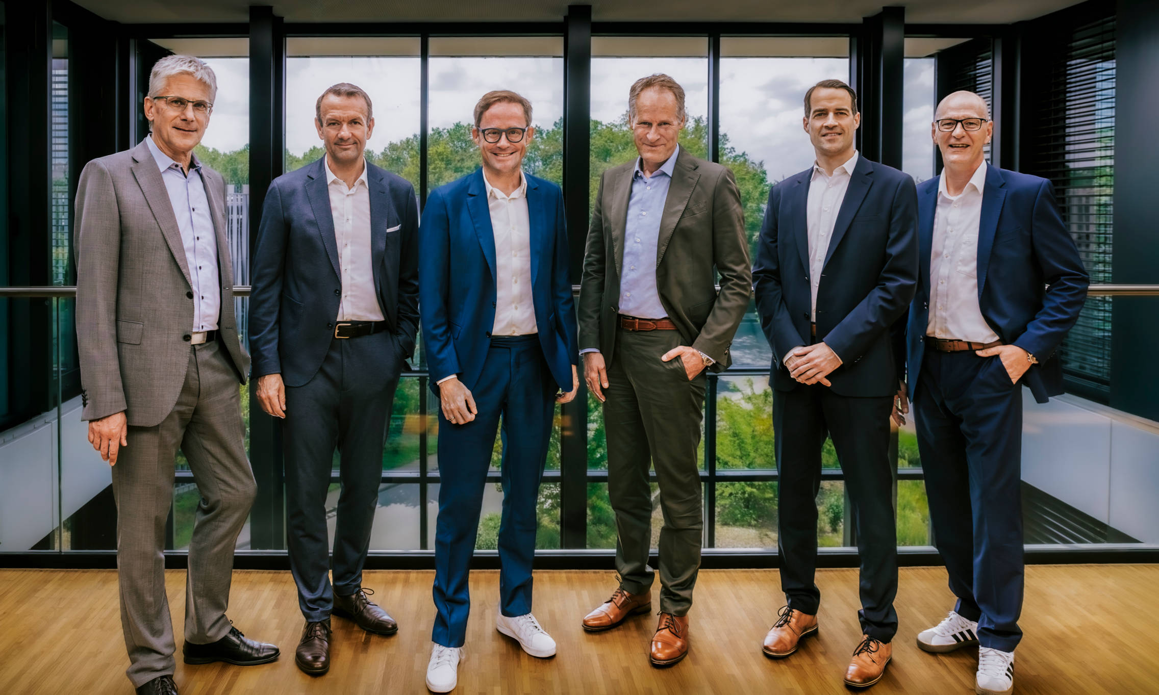 Six executives in business suits are standing in a modern office in front of a window overlooking the green countryside.
