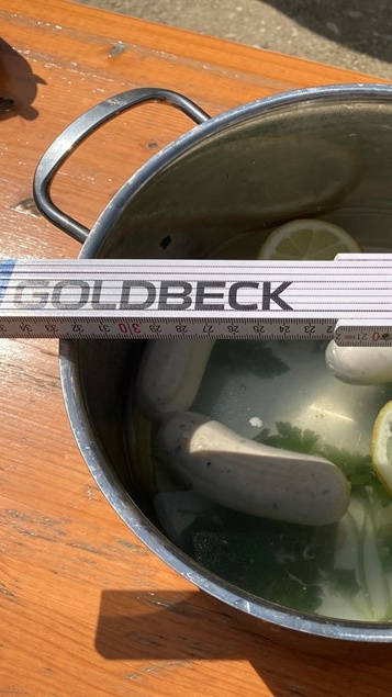 Topf mit Weißwürsten und Zitronenscheiben in Wasser auf Holztisch, daneben Zollstock.