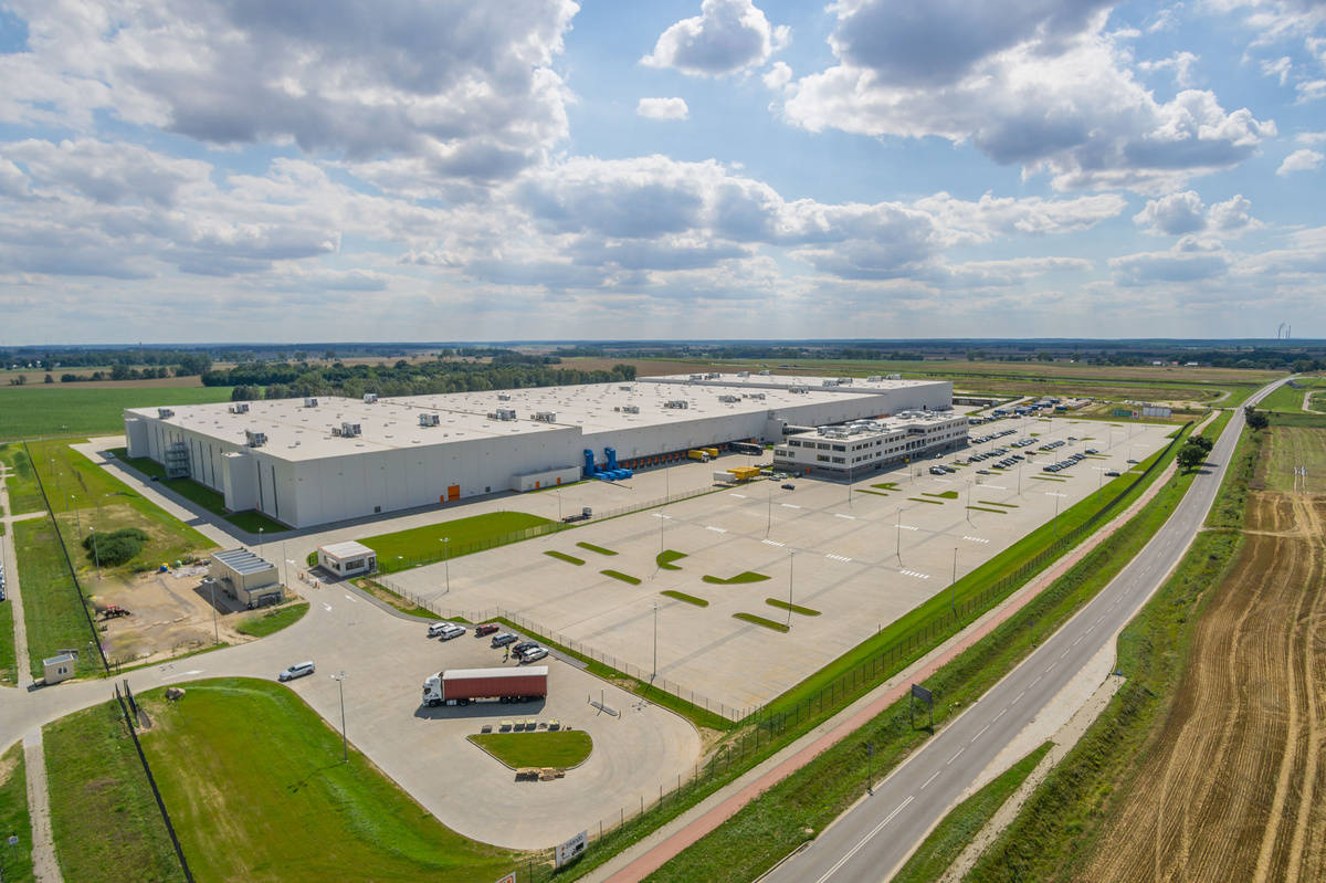 Luftaufnahme eines großen Industriegebäudes mit Parkplätzen und umliegenden Feldern unter bewölktem Himmel.