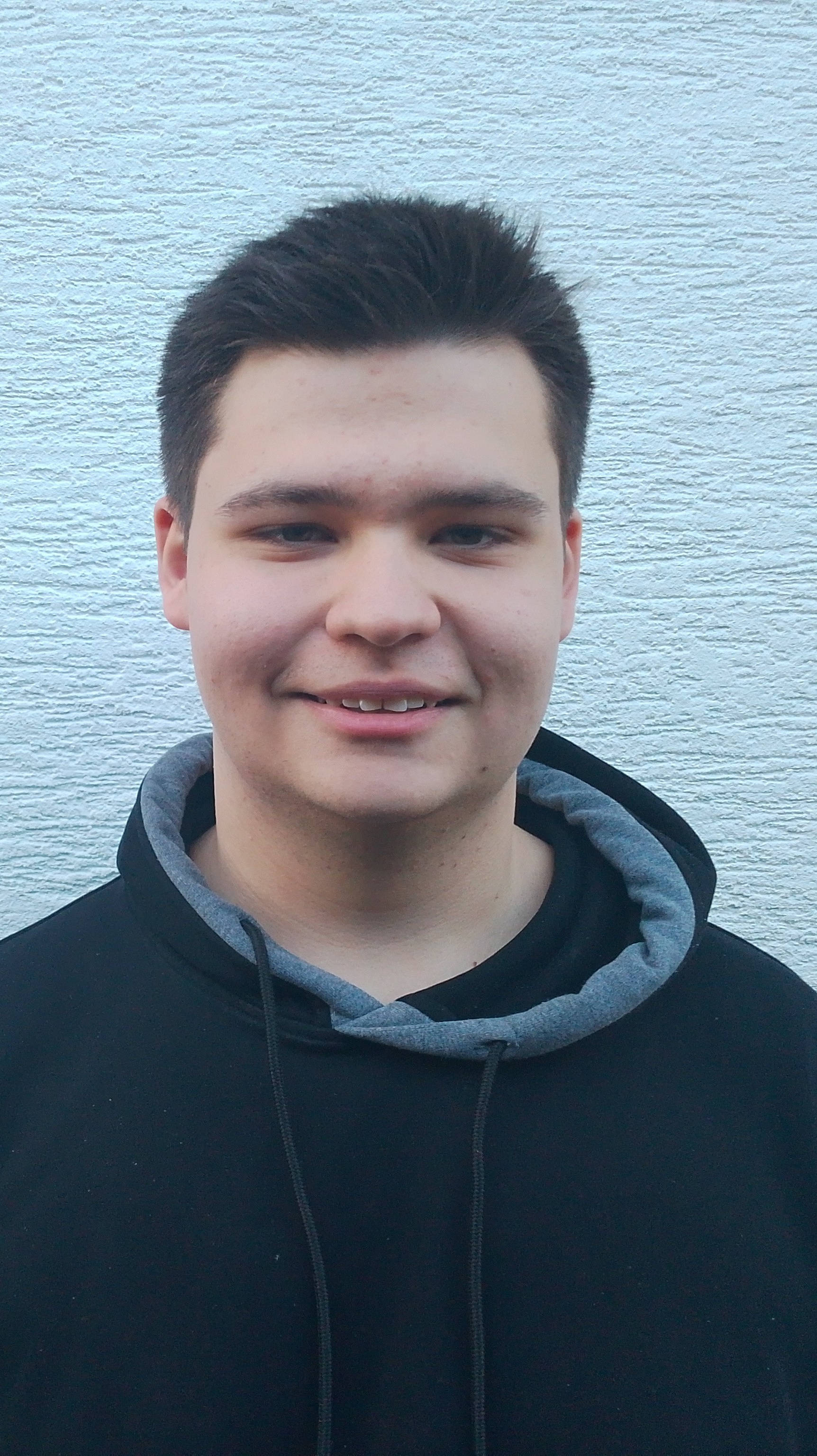 Ein junger Mann steht vor einer weißen Wand, trägt einen schwarzen Hoodie und lächelt in die Kamera.