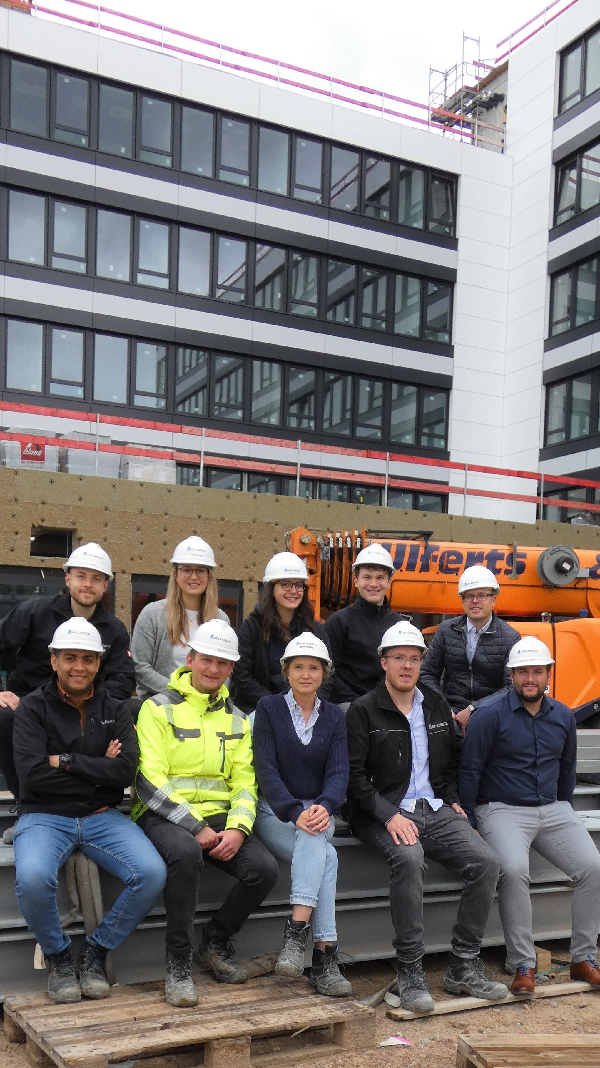 Gruppe von Bauarbeitern und Architekten mit Helmen vor einem im Bau befindlichen Bürogebäude.