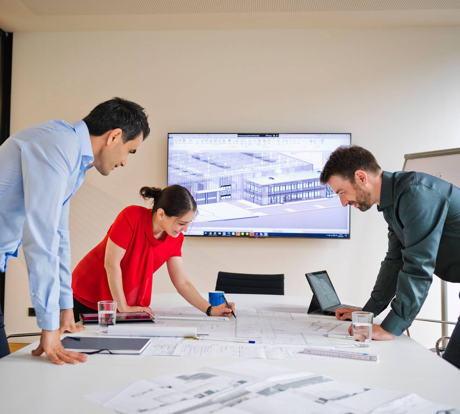 Drei Personen arbeiten in einem Besprechungsraum an Architekturplänen, mit einem digitalen Bauplan auf dem Bildschirm.